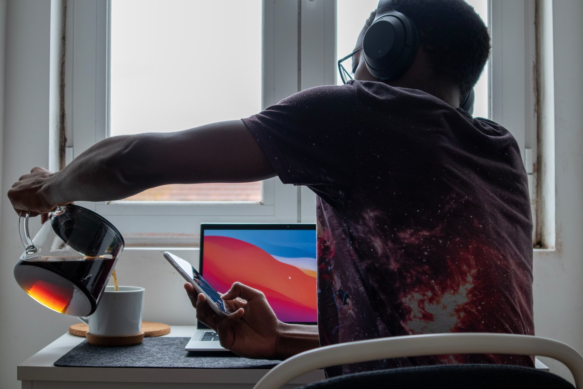 A man wearing headphones pours coffee from a glass carafe into a white mug while looking at his phone, with a laptop open on a desk.