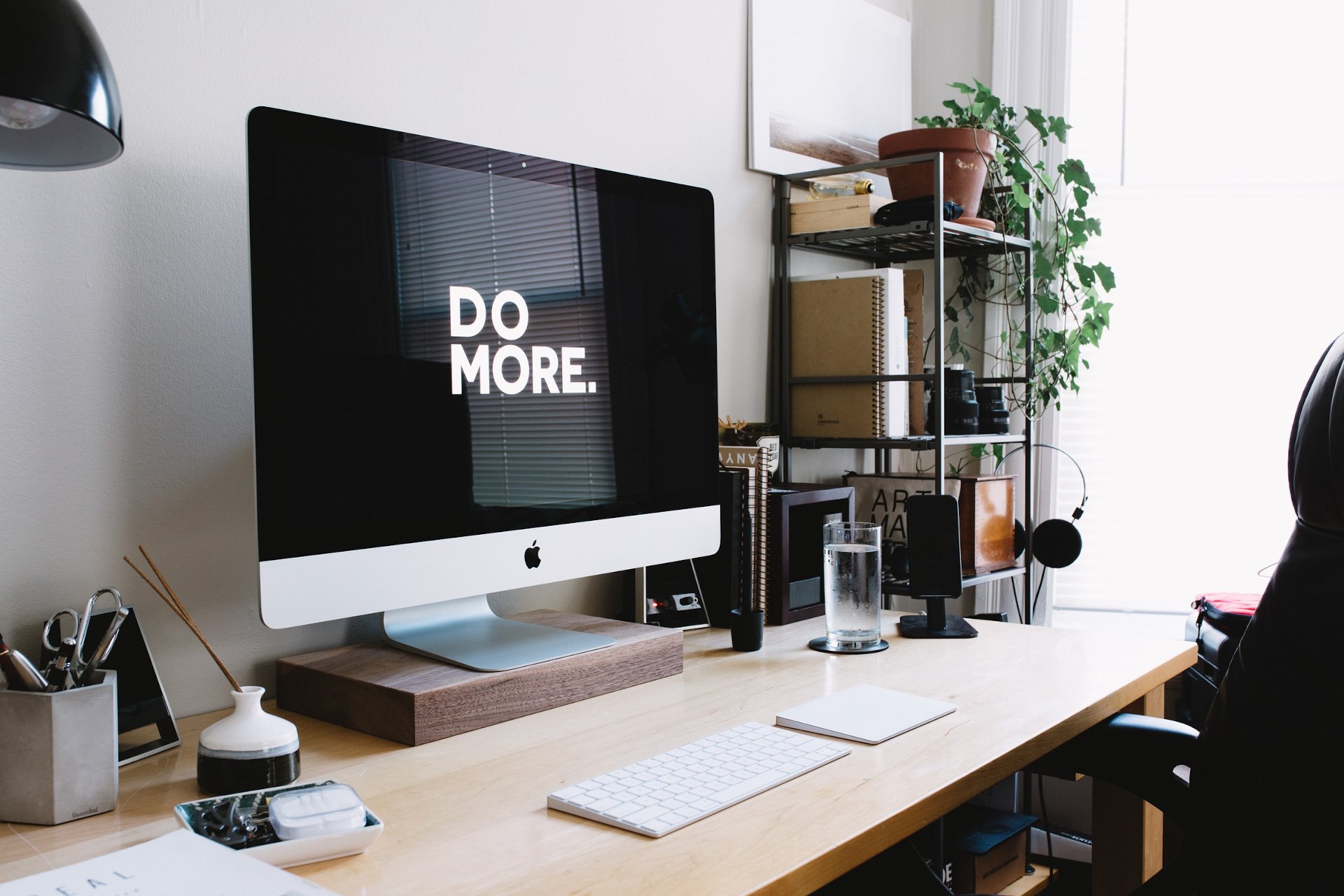 Modern home office setup with an Apple iMac displaying 'DO MORE' on a wooden desk, featuring a keyboard, mouse, headphones, and plants, promoting productivity and a clean workspace.