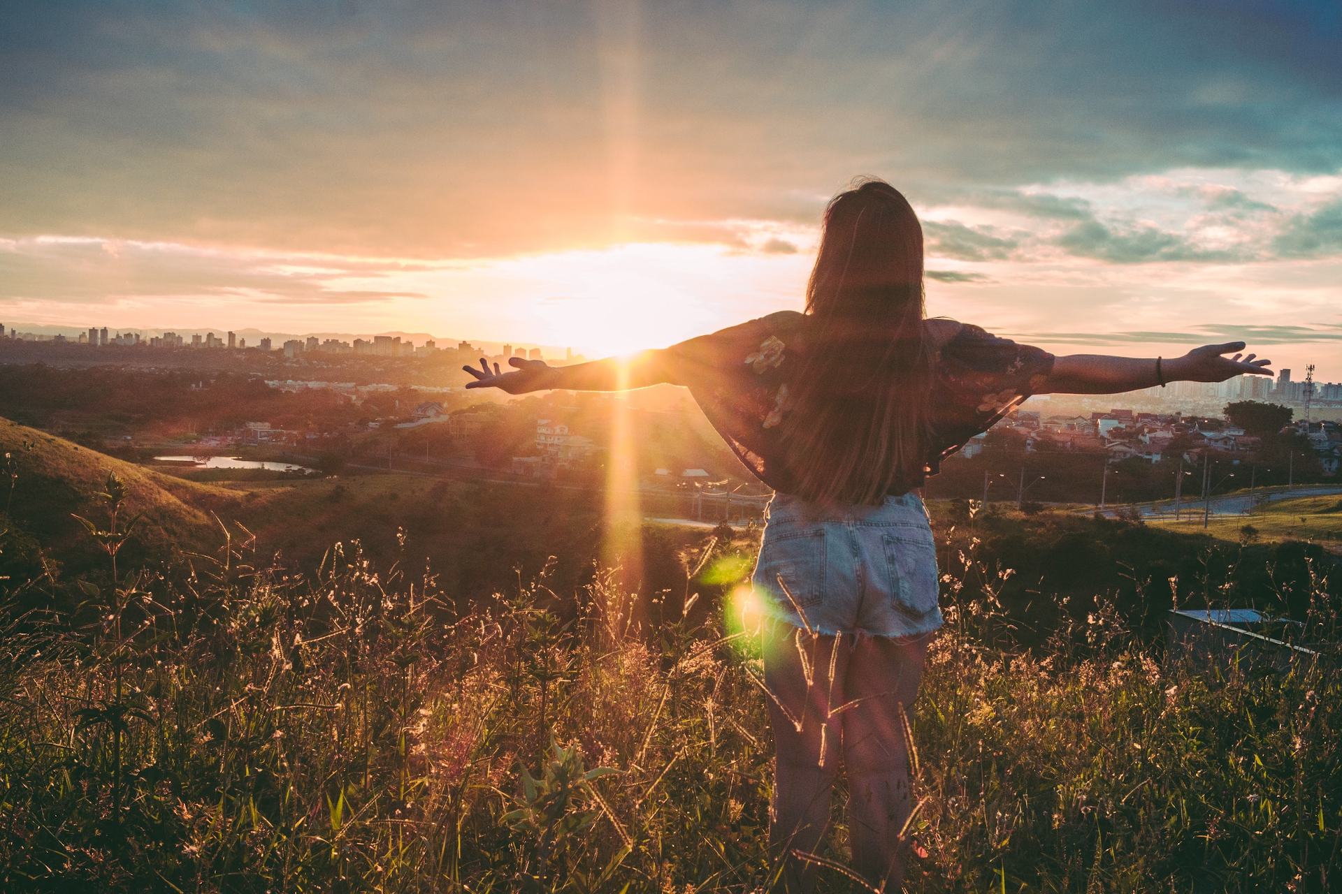 A woman stands on a hill at sunset, arms outstretched, embracing the warm glow of the sun and the city skyline.