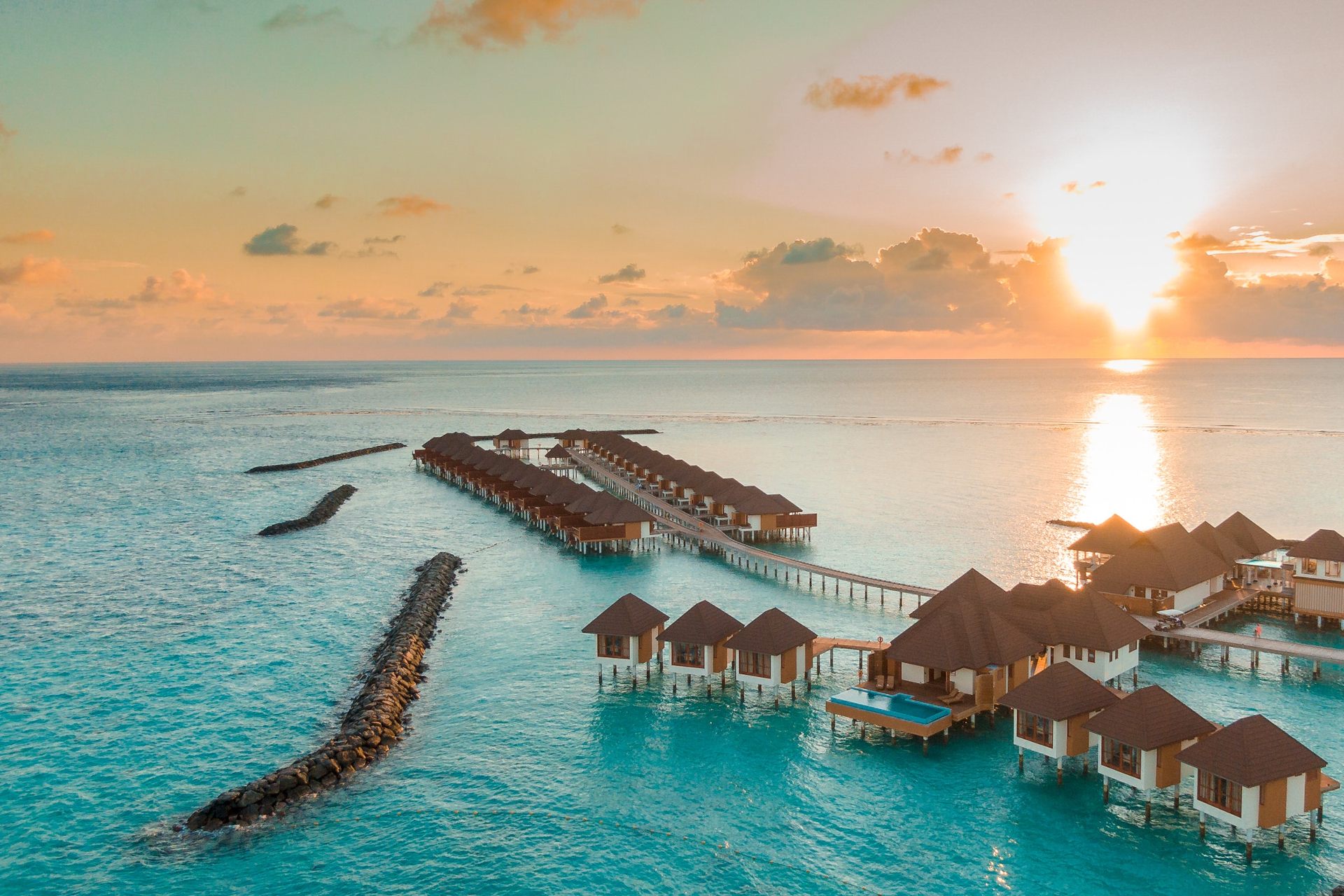 Aerial view of luxurious overwater bungalows at sunset, reflecting golden hues on the tranquil ocean waters.